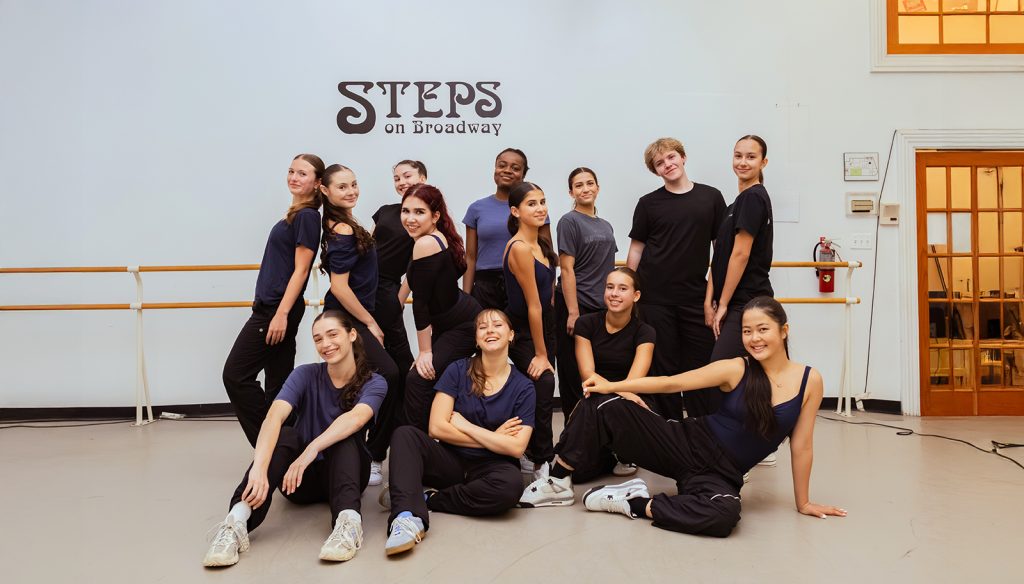 A large group of Steps Academy dancers posed in front of the barre and Steps logo at Steps Academy