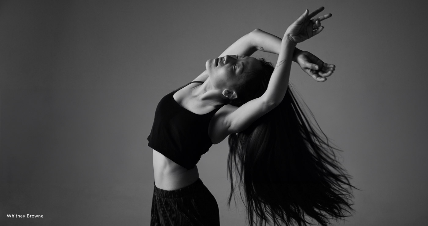 Black and white photo of a single female dancer seen from the hips up, head thrown back and arms up with long flowing dark hair