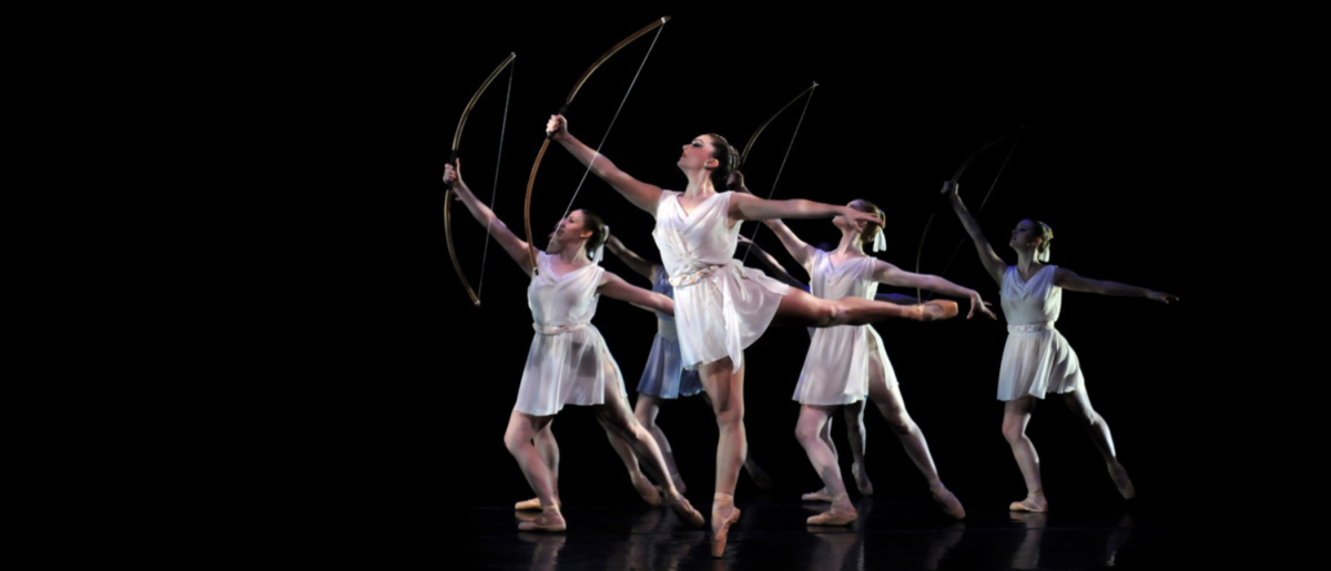 University of Hartford dancers in white on a black background with archery bows and wearing toga-like dresses