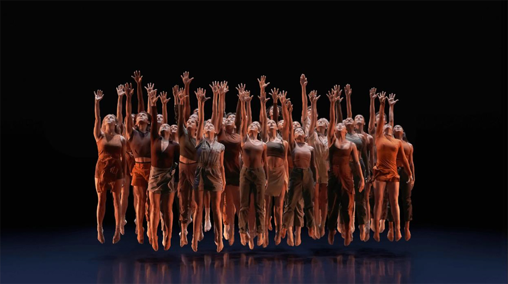 A Large group of dance tightly grouped under the spotlight on a dark stage, each with one arm raised and fingers spread towards the sky